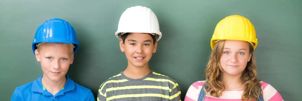 © pictworks / Adobe Stock 3 lächelnde Kinder mit Bauhelmen auf dem Kopf.